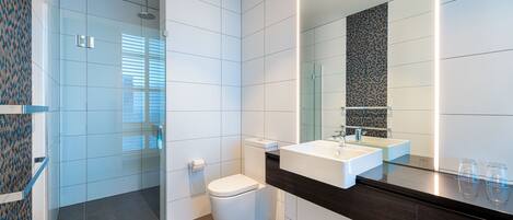 Un baño moderno con cabina de ducha de vidrio, inodoro blanco, tocador de madera oscura con un espejo grande y lavamanos blanco.