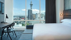 A hotel room with a large window offering a city view, a bed, a desk with a chair, and a lamp. - Four Points by Sheraton Auckland (Auckland)