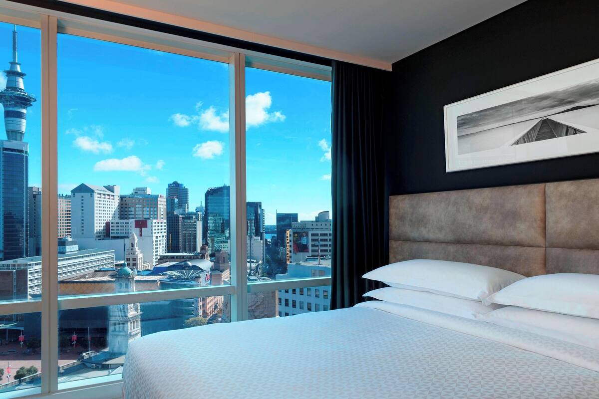 A hotel room with a large window offering a city view, a bed with white linens, and a framed picture on the wall.