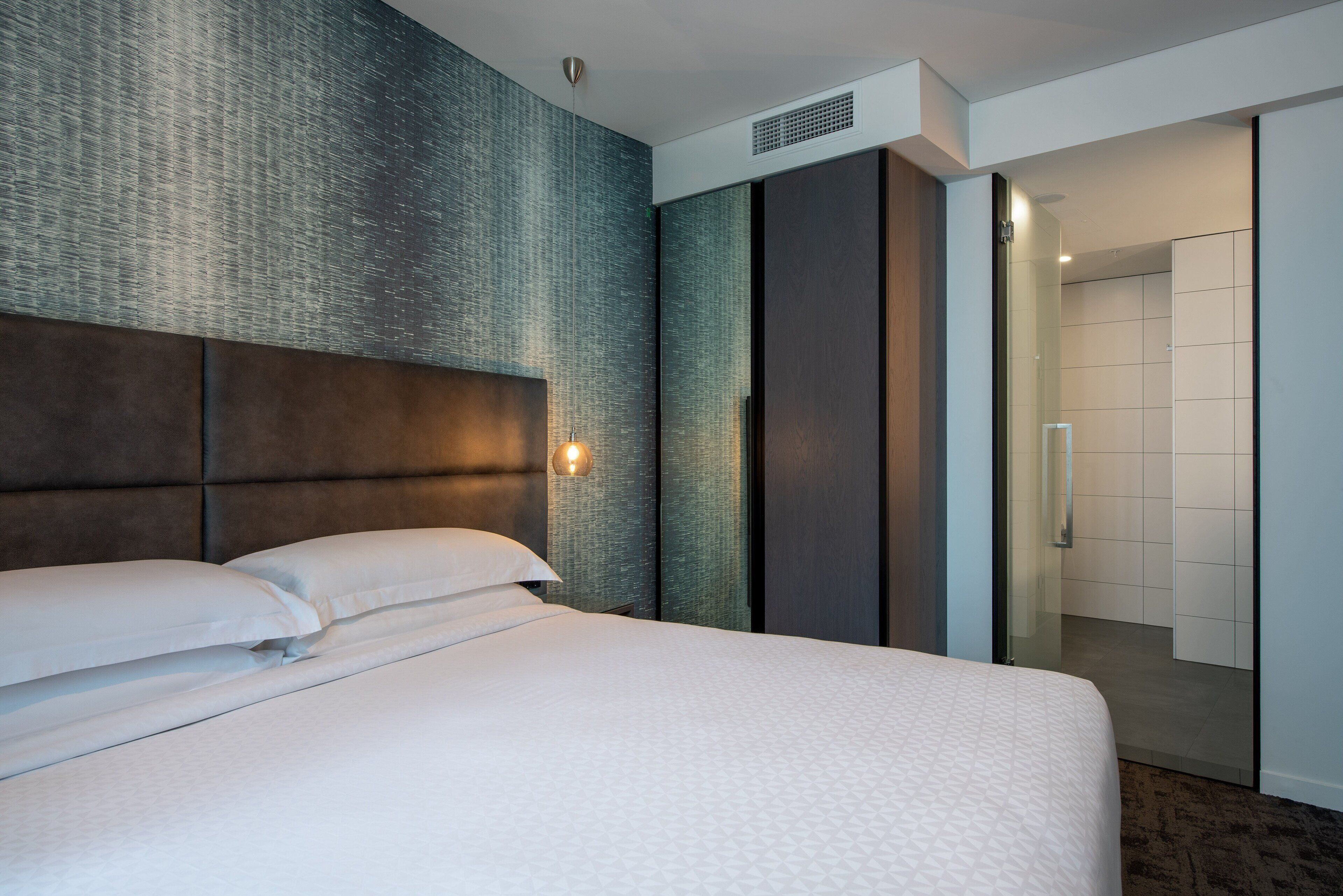 A modern hotel room with a large bed, a textured wall, and a sliding door leading to a bathroom.