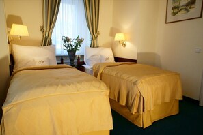 A hotel room with two beds, each with a matching headboard and pillow, a bedside table with a lamp, a window with curtains, and a painting on the wall. - Hotel Mignon (Karlovy Vary)