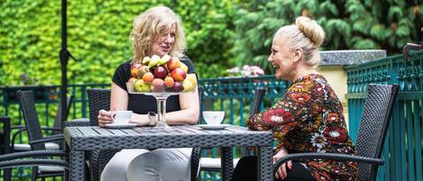 Two people seated at an outdoor table with a fruit bowl, surrounded by wicker chairs and a green fence.