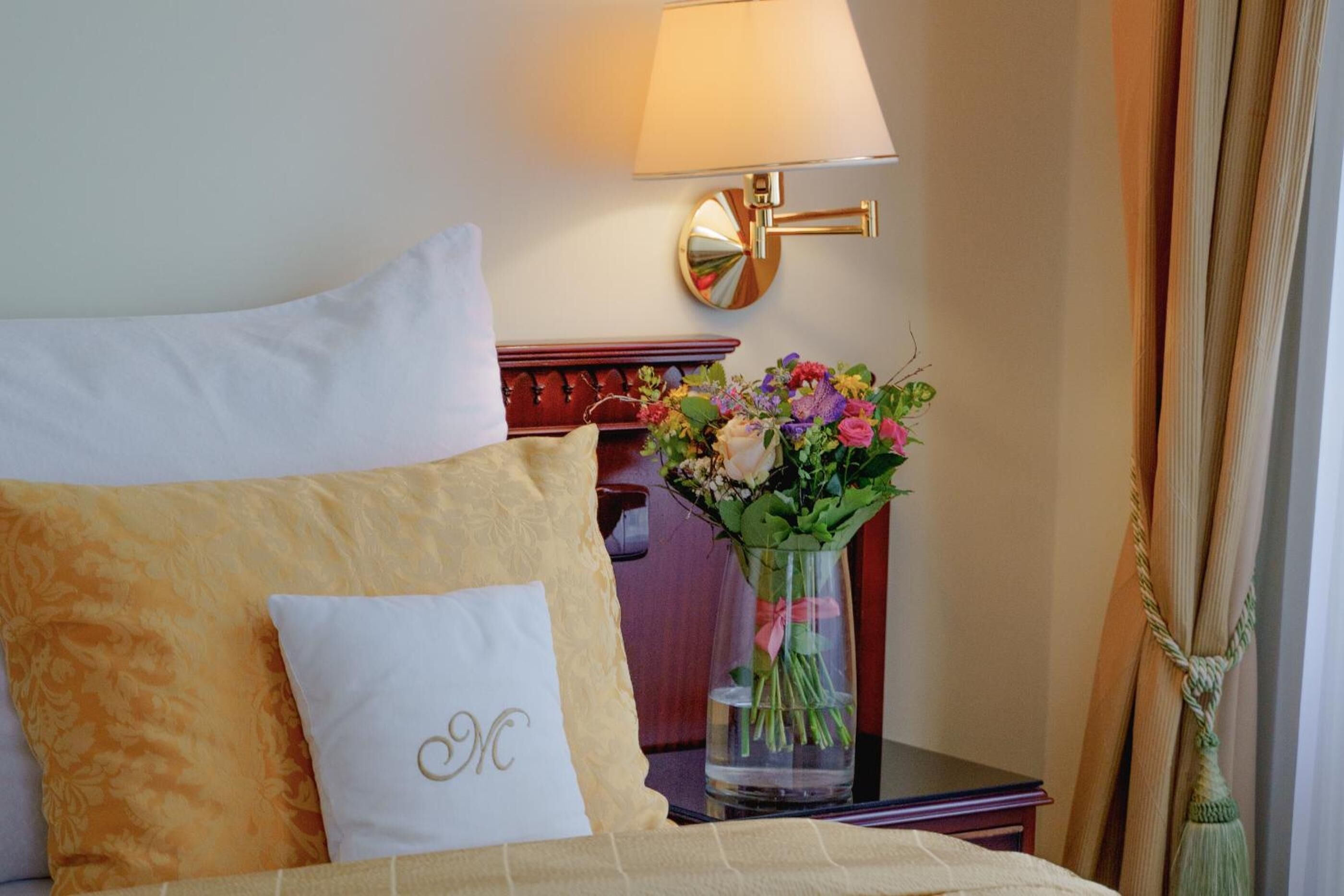Una camera d'albergo con un letto, un comodino con una lampada, un vaso di fiori e uno specchio decorativo.