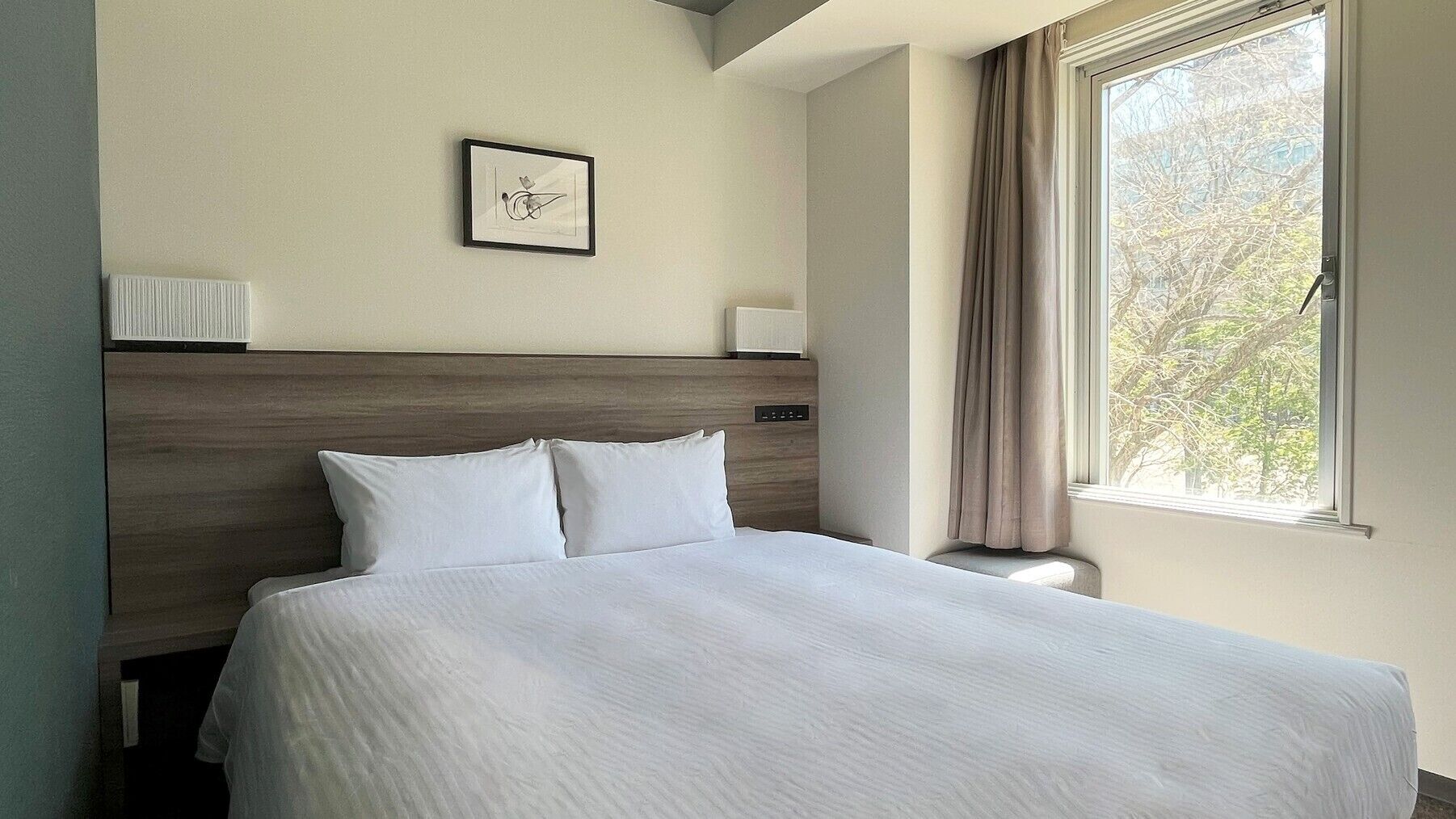 Une chambre d’hôtel avec un grand lit, une tête de lit, une table de chevet, une lampe, un tableau encadré au mur et une fenêtre avec des rideaux.