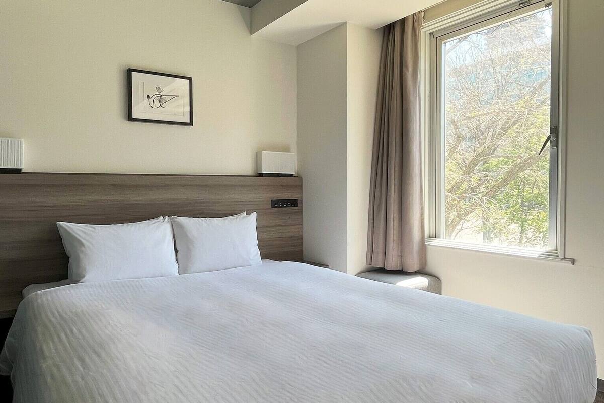 A hotel room with a large bed, a headboard, a bedside table, a lamp, a framed picture on the wall, and a window with curtains.
