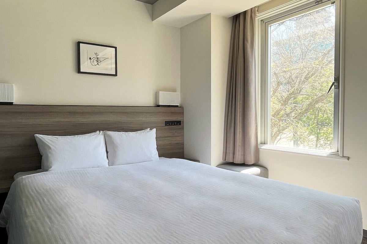 Habitación de hotel con una cama grande, cabecero, mesita de noche, lámpara, un cuadro enmarcado en la pared y una ventana con cortinas.