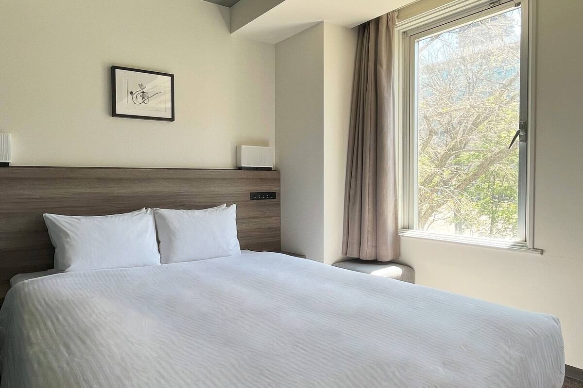 A hotel room with a large bed, a headboard, a bedside table, a lamp, a framed picture on the wall, and a window with curtains.