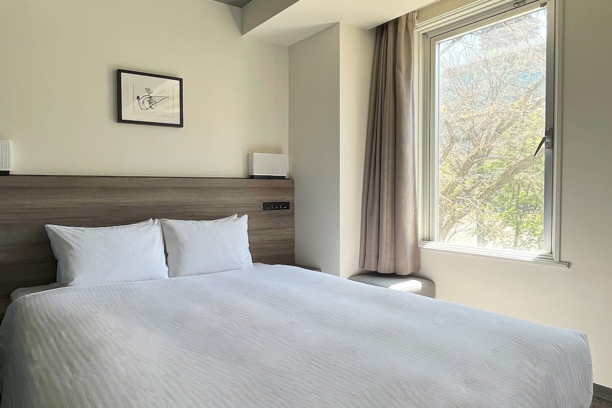 Une chambre d’hôtel avec un grand lit, une tête de lit, une table de chevet, une lampe, un tableau encadré au mur et une fenêtre avec des rideaux.