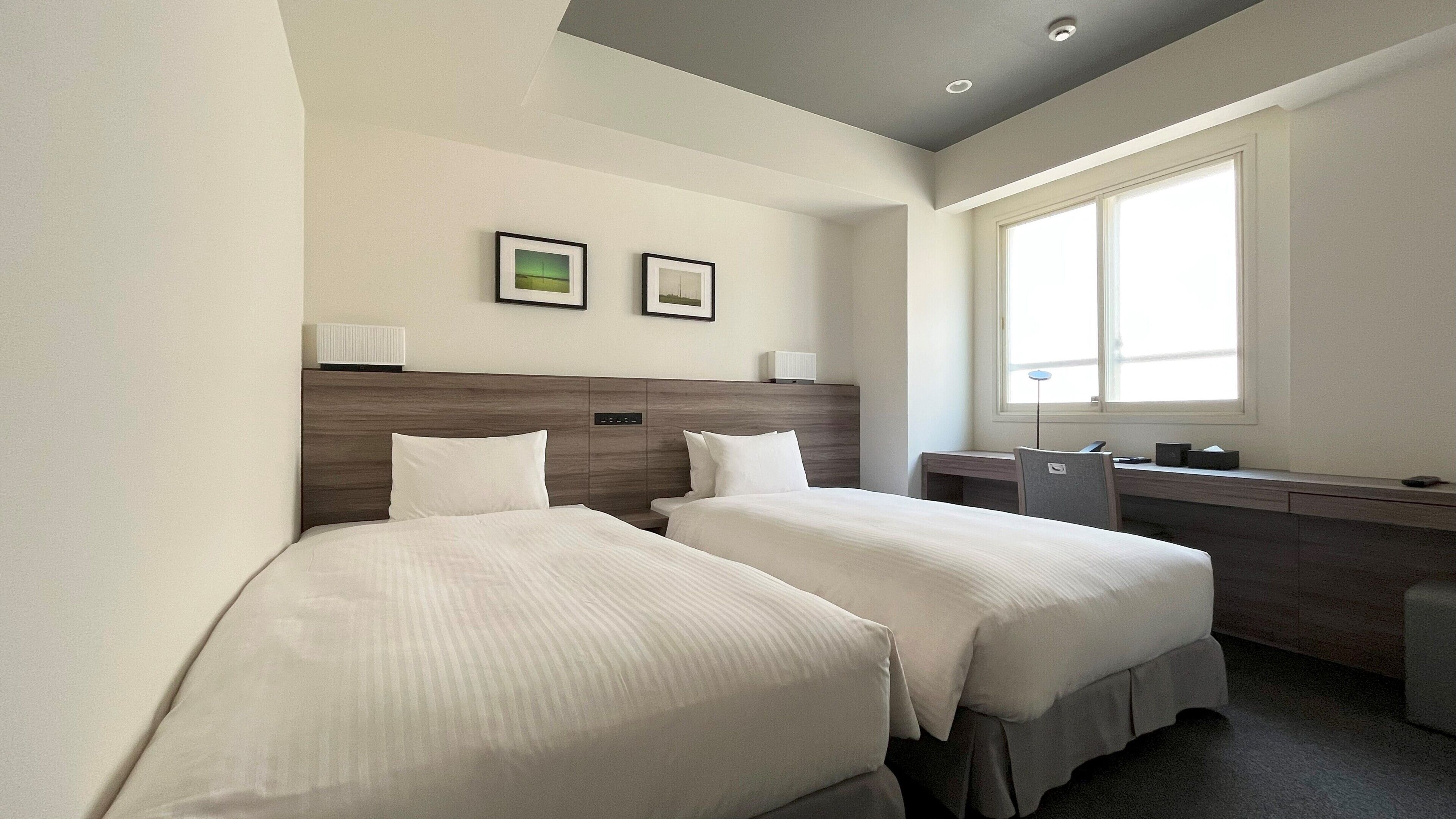 Een hotelkamer met twee enkele bedden, een bureau, een stoel, een raam en twee ingelijste foto's aan de muur.