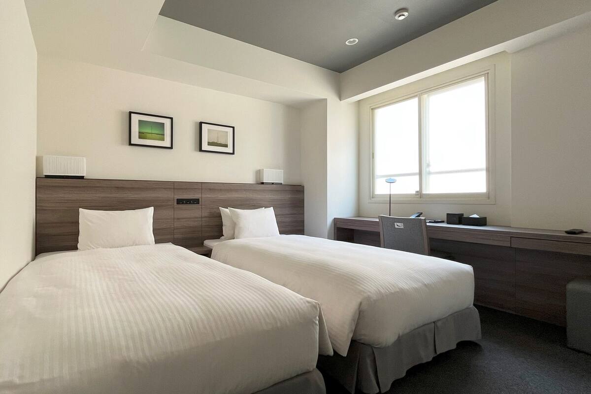 A hotel room with two single beds, a desk, a chair, a window, and two framed pictures on the wall.