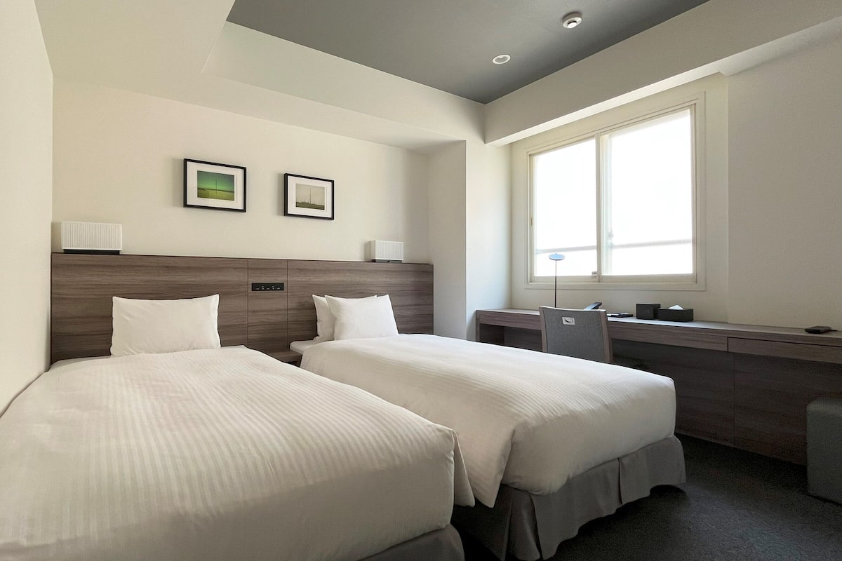 Une chambre d’hôtel avec deux lits simples, un bureau, une chaise, une fenêtre et deux tableaux encadrés au mur.