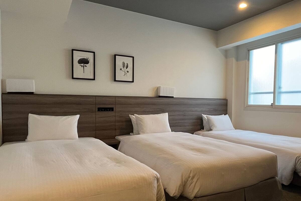 A hotel room with two single beds, a headboard, bedside lamps, and a window with natural light.