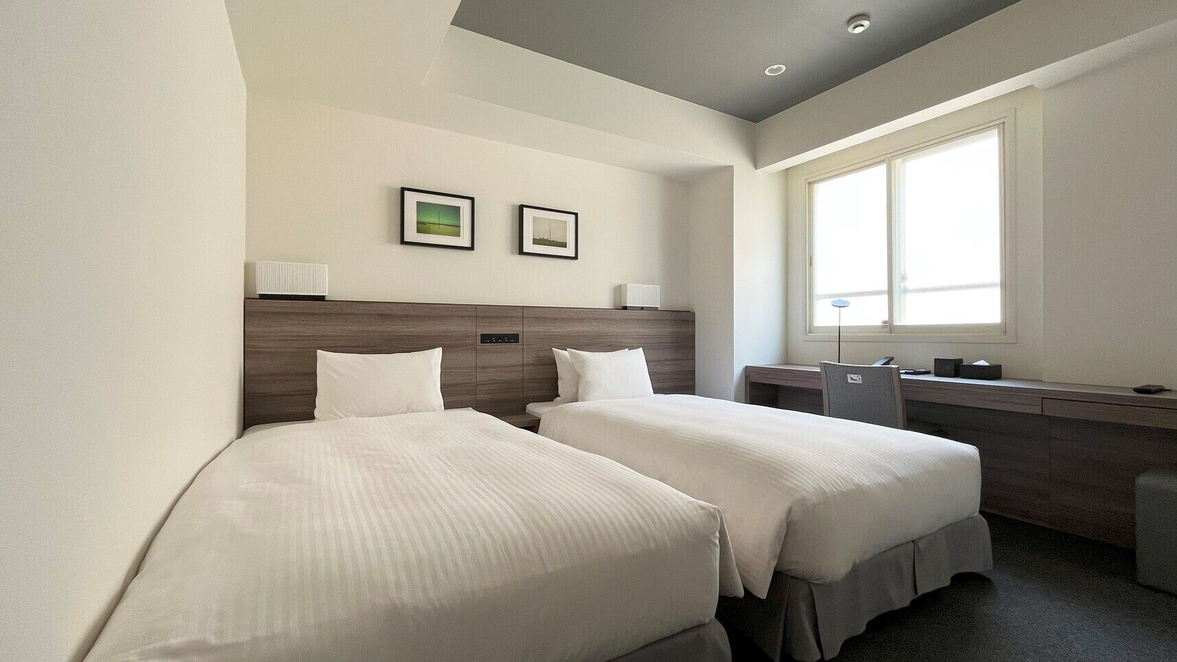 Une chambre d’hôtel avec deux lits, un bureau, une chaise, une fenêtre et deux tableaux encadrés au mur.