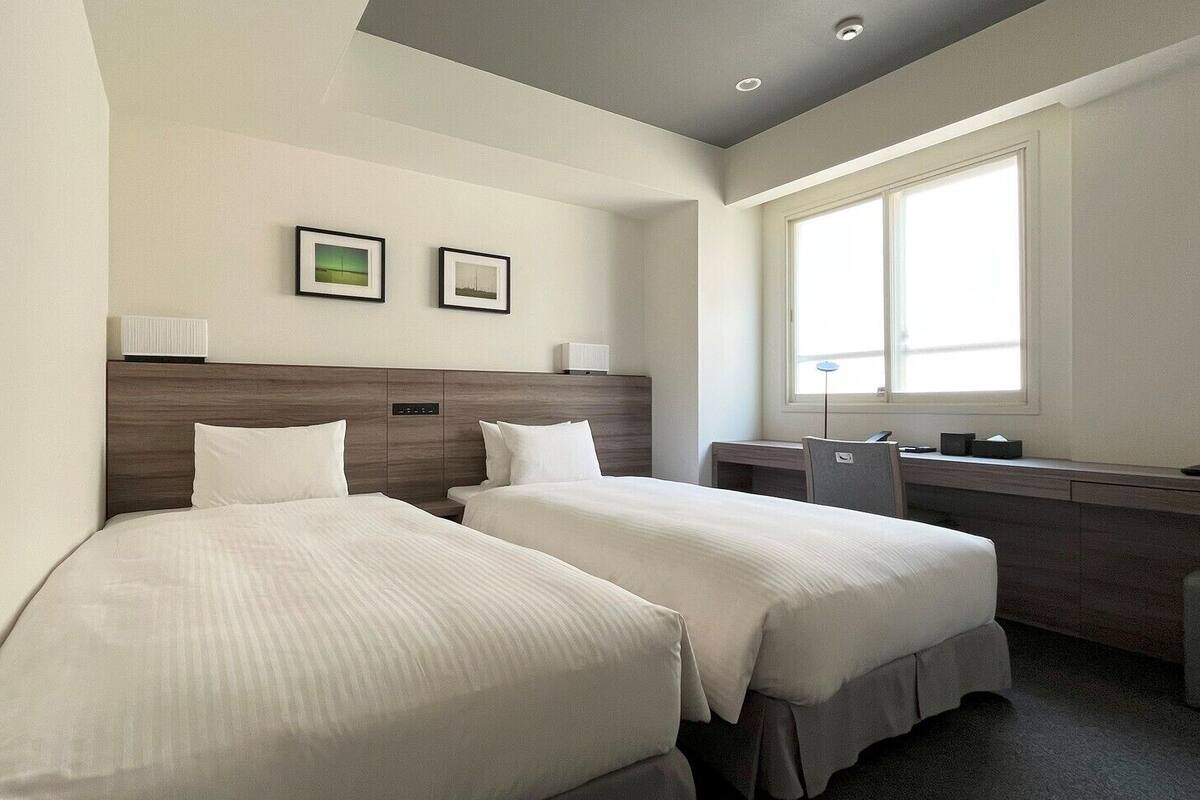 A hotel room with two beds, a desk, a chair, a window, and two framed pictures on the wall.