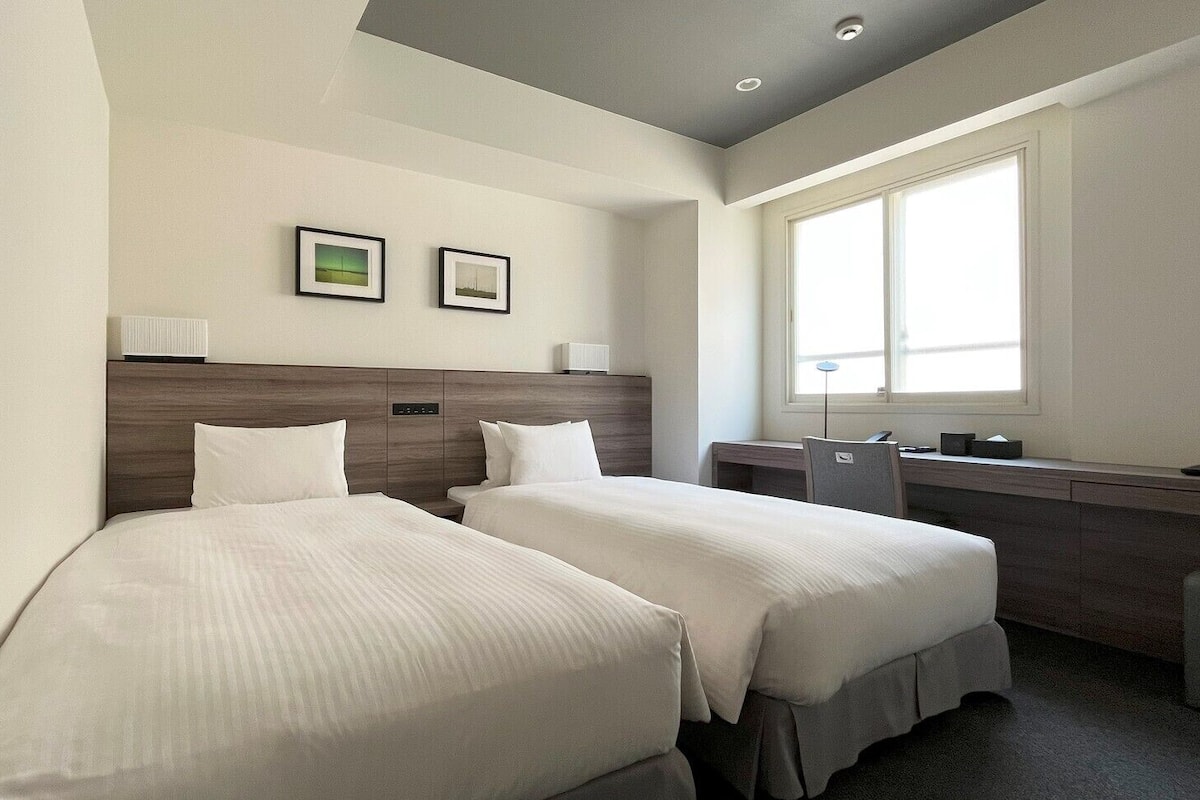 Habitación de hotel con dos camas, un escritorio, una silla, una ventana y dos cuadros en la pared.