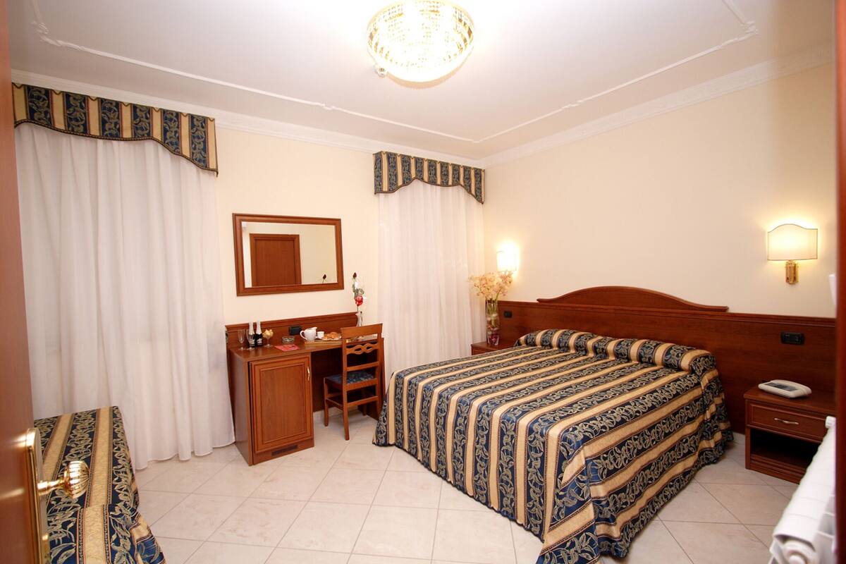 A hotel room with a large bed, a desk with a chair, a mirror, a bedside table with a lamp, and curtains.