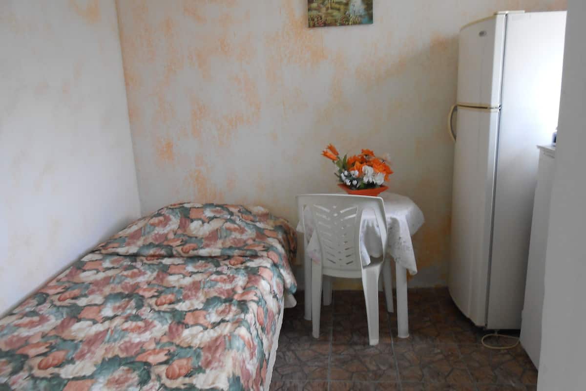 Ein kleines Zimmer mit einem Bett, einem Tisch mit Blumenvase, einem Stuhl und einem Kühlschrank.