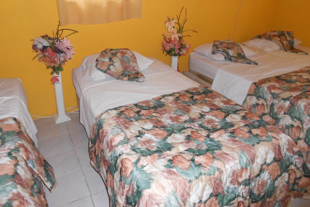 Ein Hotelzimmer mit drei Betten, jedes mit floralem Überwurf und passenden Kissen. Auf den Nachttischen stehen jeweils Blumenarrangements.