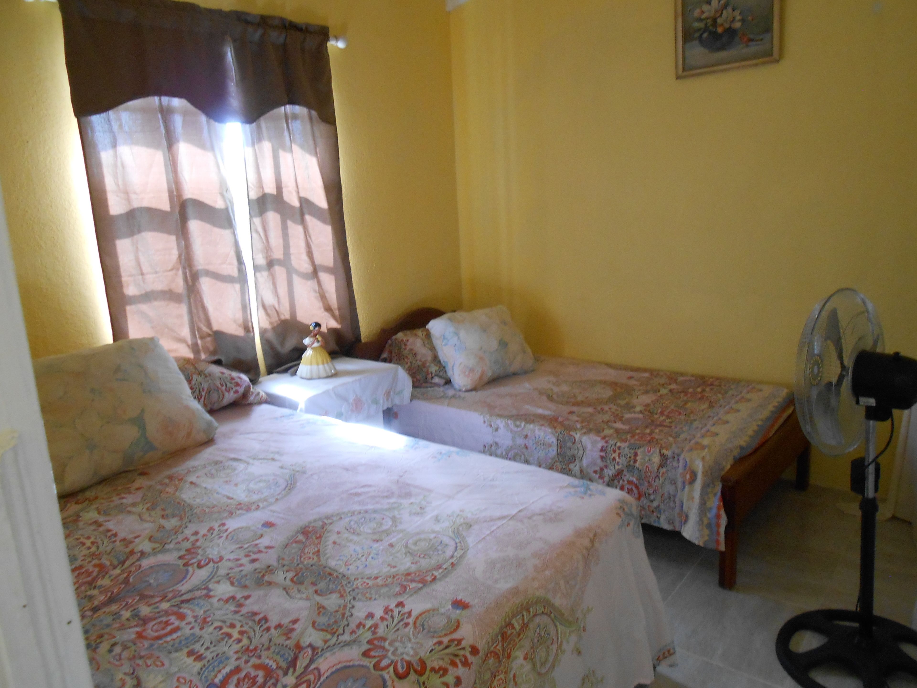 A room with two beds, a table with a figurine, a fan, and a painting on the wall.