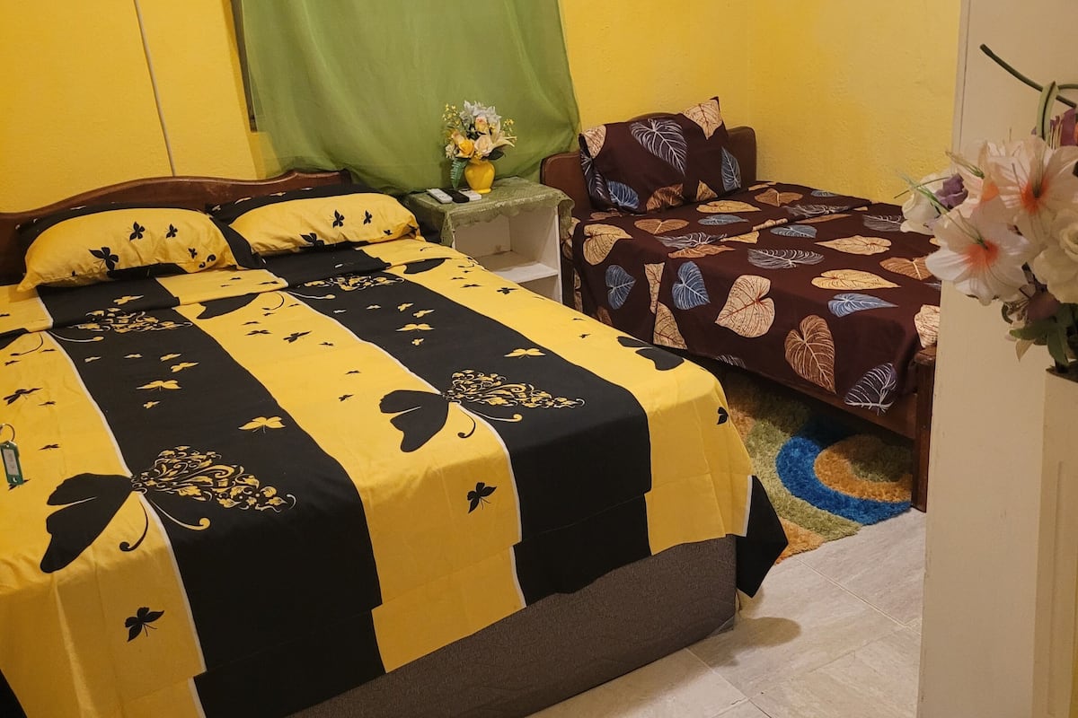 Ein Zimmer mit einem Bett, einem Sofa, einem kleinen Tisch mit Blumen und einem grünen Vorhang.