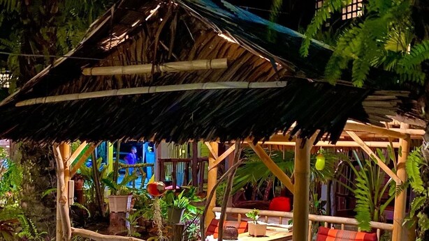 Un espacio al aire libre con techo de paja, con varios bancos y cojines, rodeado de abundante follaje y plantas en macetas.