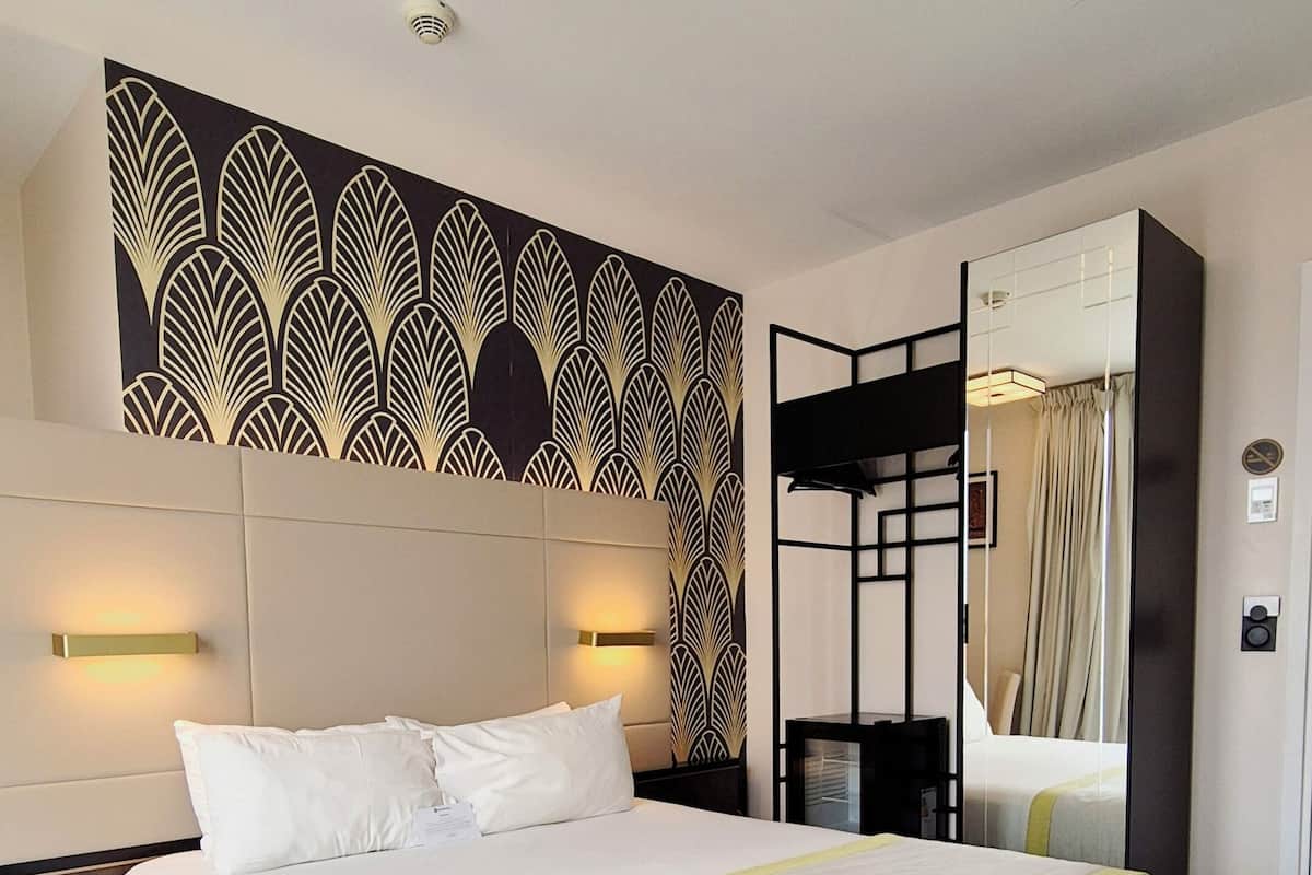 A modern hotel room with a large bed, a decorative wall panel, a mirror, and a ceiling light fixture.