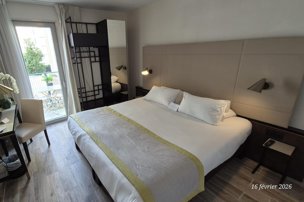 A modern hotel room with a large bed, a desk with a chair, a desk lamp, and a view of the outdoors through a sliding door.