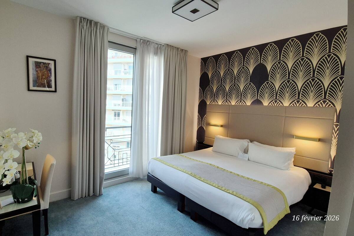 A hotel room with a large bed, a desk with a chair, a vase of white flowers, and a window with curtains.