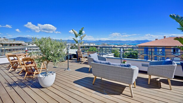 Um deck de madeira com móveis para área externa, um vaso de planta e vista para o mar e edifícios.