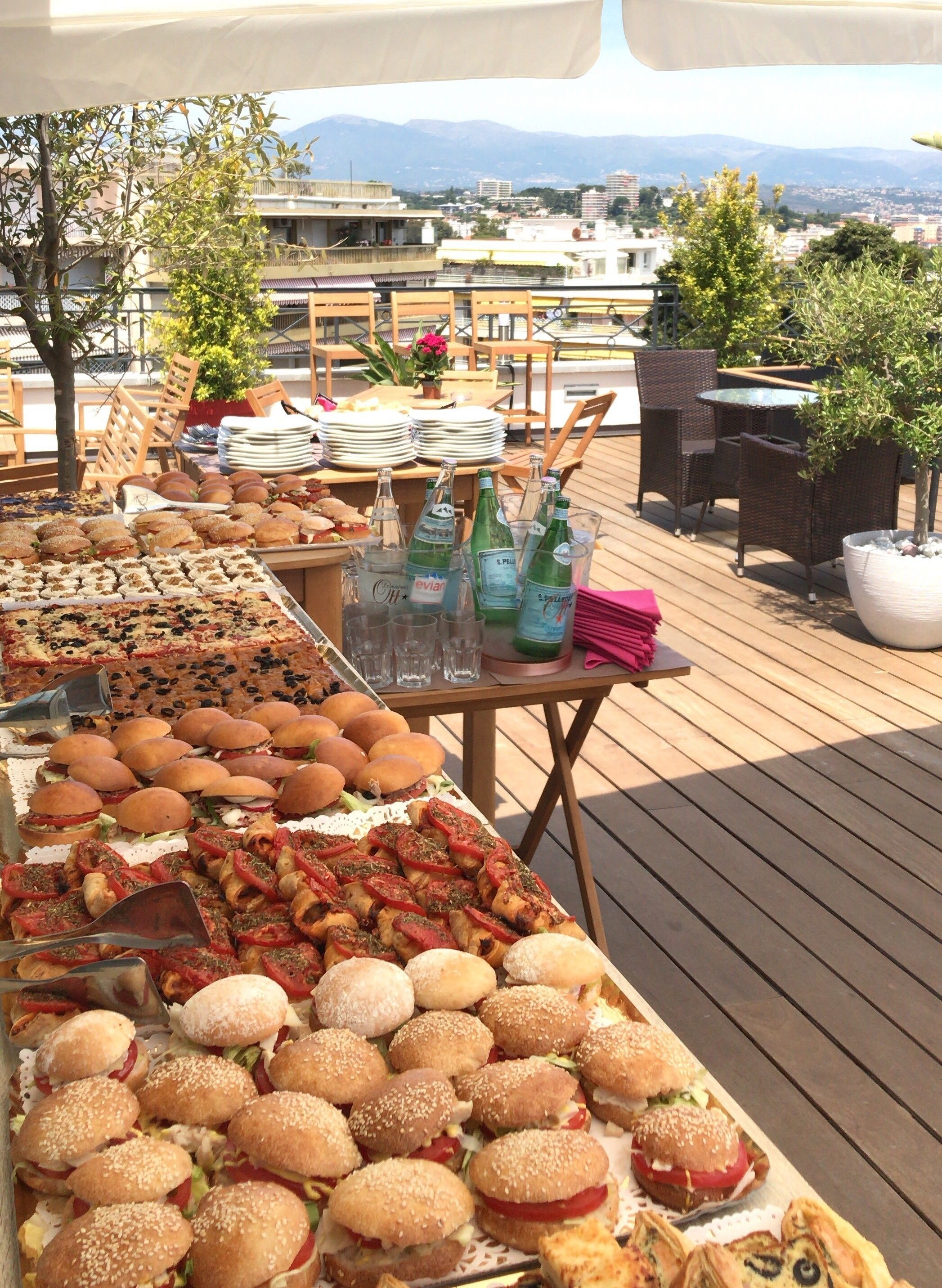 Un bufé al aire libre con diversas opciones de comida y bebidas en una terraza de madera, con vista a edificios y montañas al fondo.