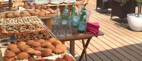 Un bufé al aire libre con diversas opciones de comida y bebidas en una terraza de madera, con vista a edificios y montañas al fondo.