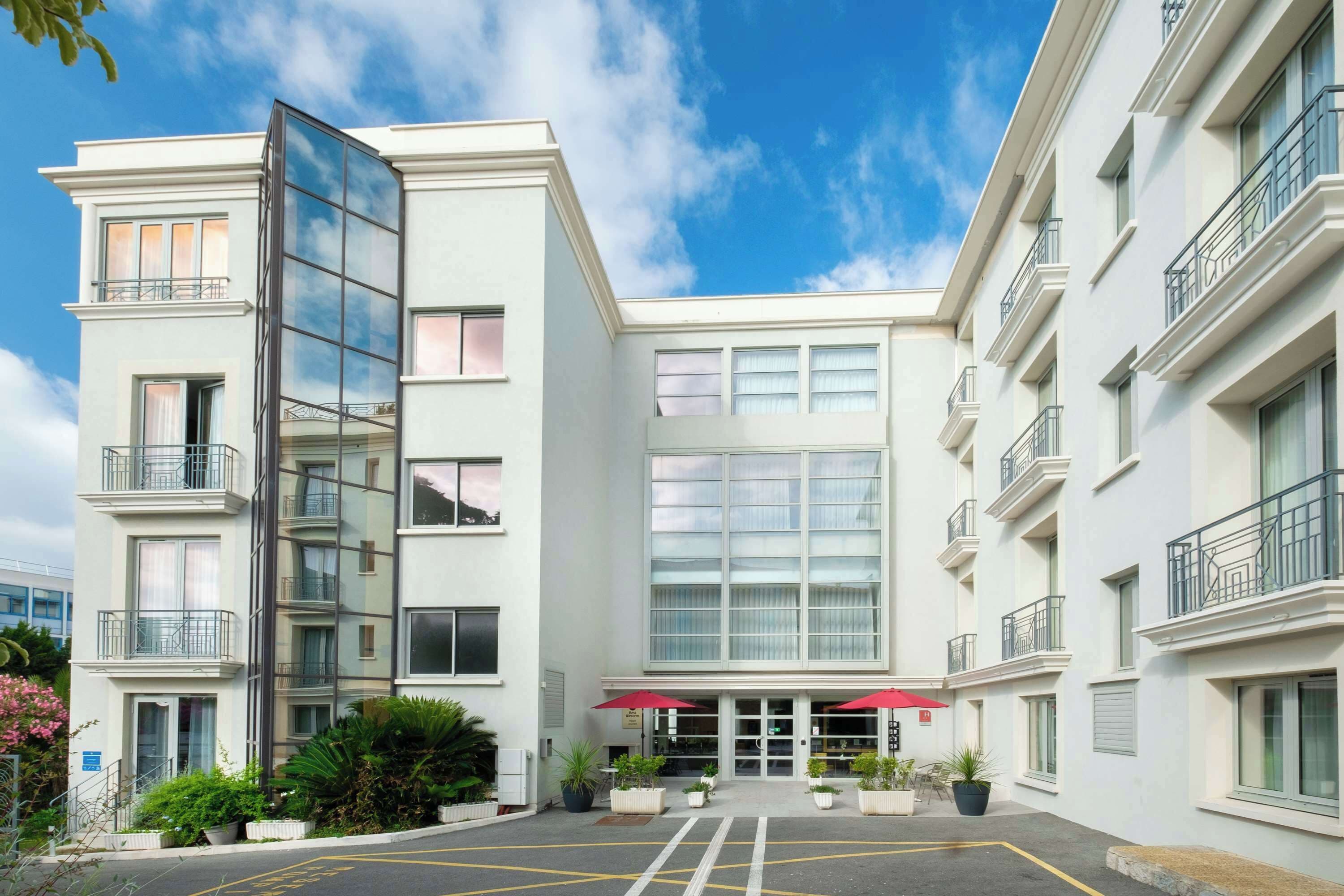 A modern building with a glass facade, multiple balconies, and a central entrance with red umbrellas.