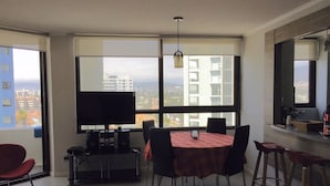 Una sala de estar moderna con un ventanal grande, un televisor de pantalla plana, una zona de comedor con una mesa y sillas, y una encimera de cocina con una cafetera.