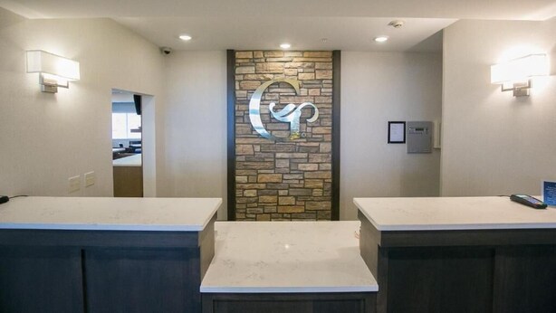 A reception area with a stone wall and a large illuminated 'G' logo. There are two reception counters and a door leading to another room. - GrandStay Cannon Falls (Cannon Falls)