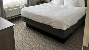 A hotel room with a large bed, a bedside table, a lamp, a window with curtains, and a wall-mounted air conditioning unit.