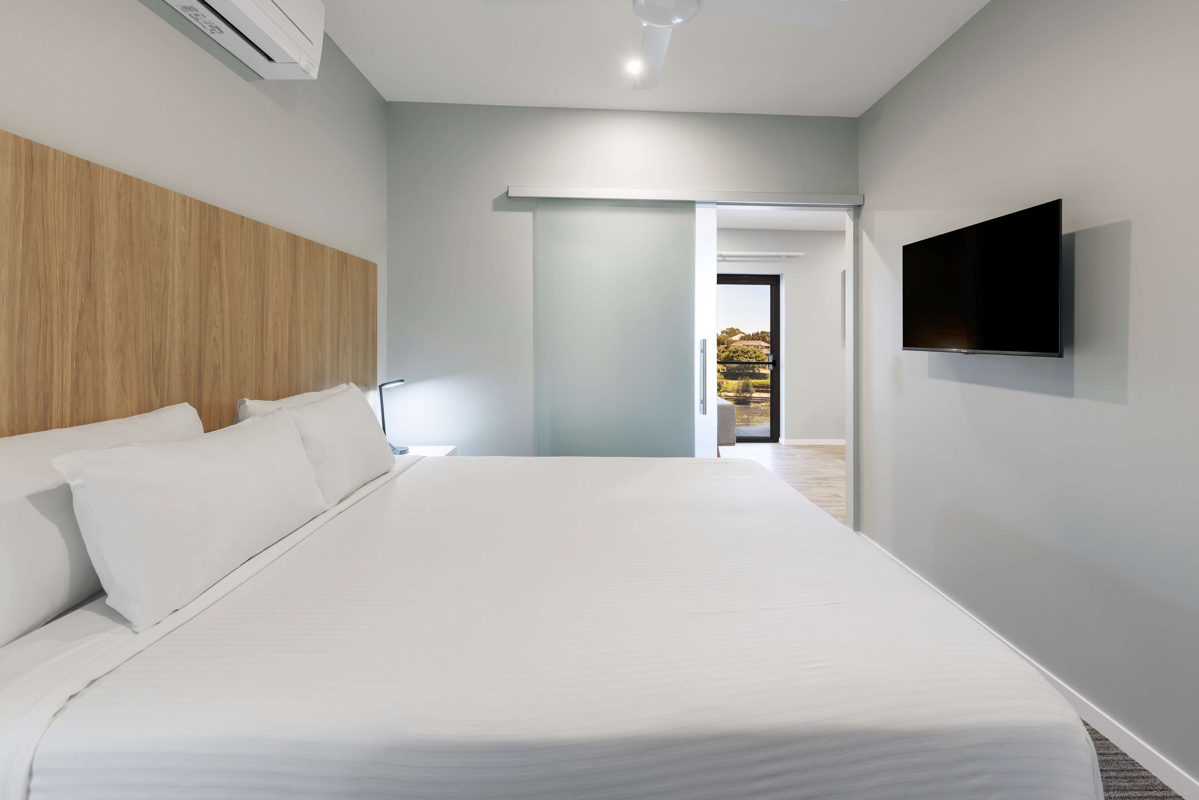 A modern bedroom with a large bed, a mounted TV, and a view of the outdoors through a door.