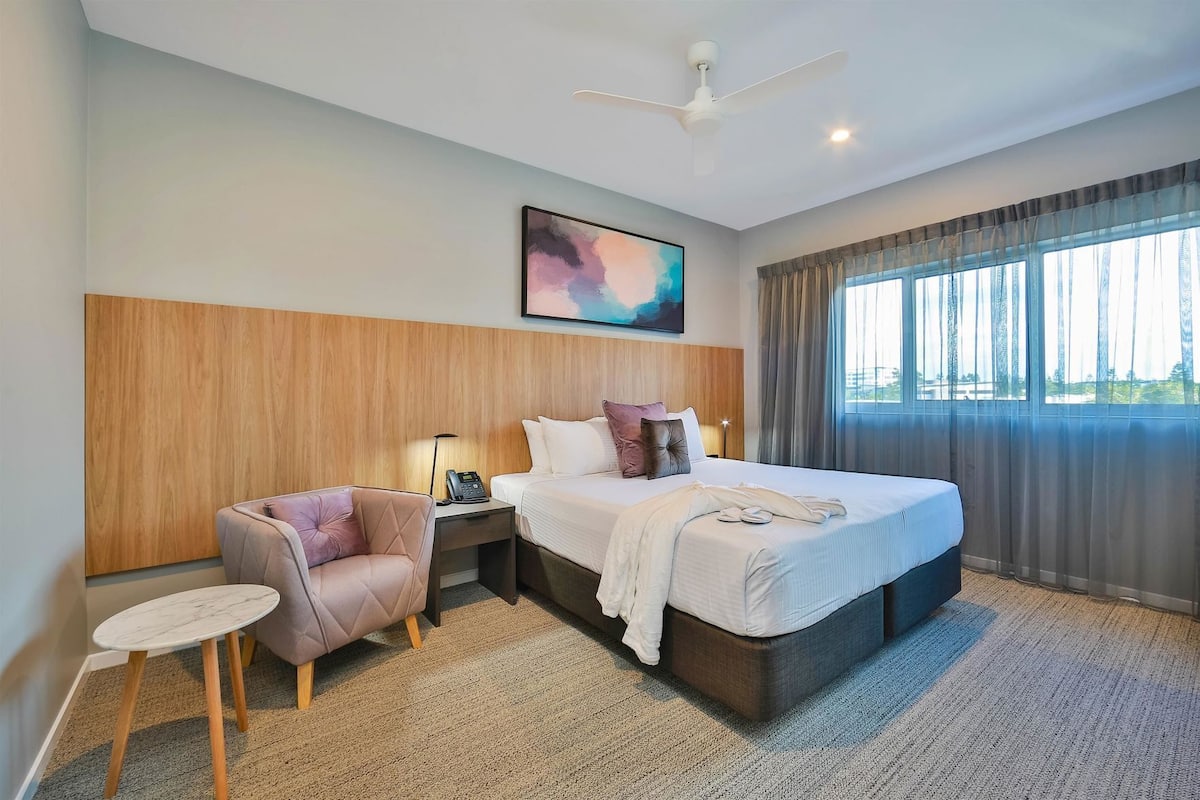 Une chambre d’hôtel avec un lit, une chaise, une table de chevet, un ventilateur de plafond, un tableau et une fenêtre avec des rideaux.