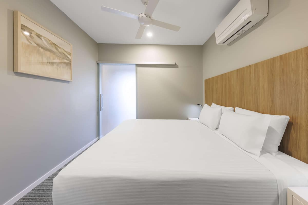 Une chambre d’hôtel avec un grand lit, un ventilateur de plafond, un appareil de climatisation, une tête de lit en bois et une porte donnant sur une autre pièce.