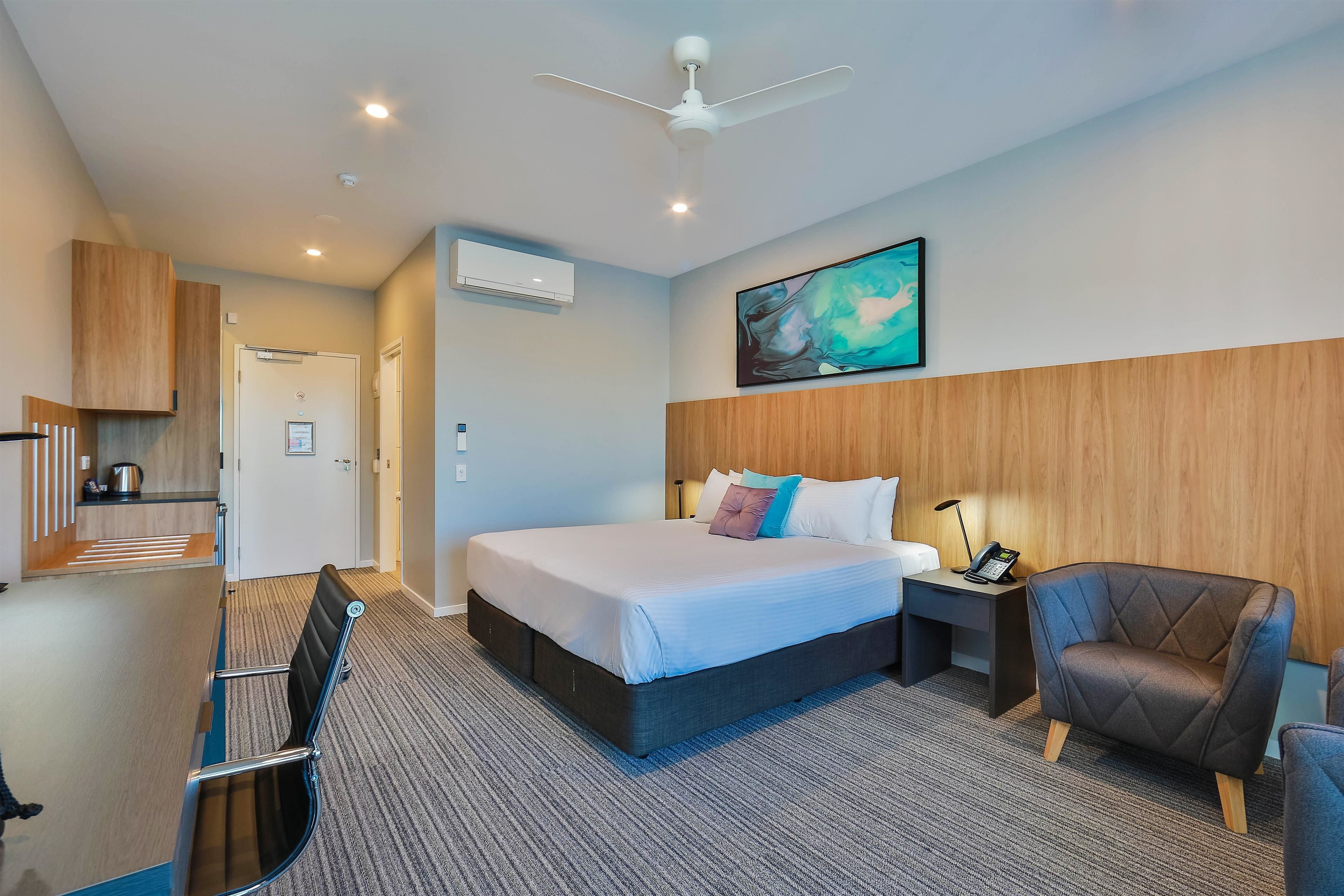 A modern hotel room with a large bed, a desk with a phone, a chair, a ceiling fan, and a painting on the wall.