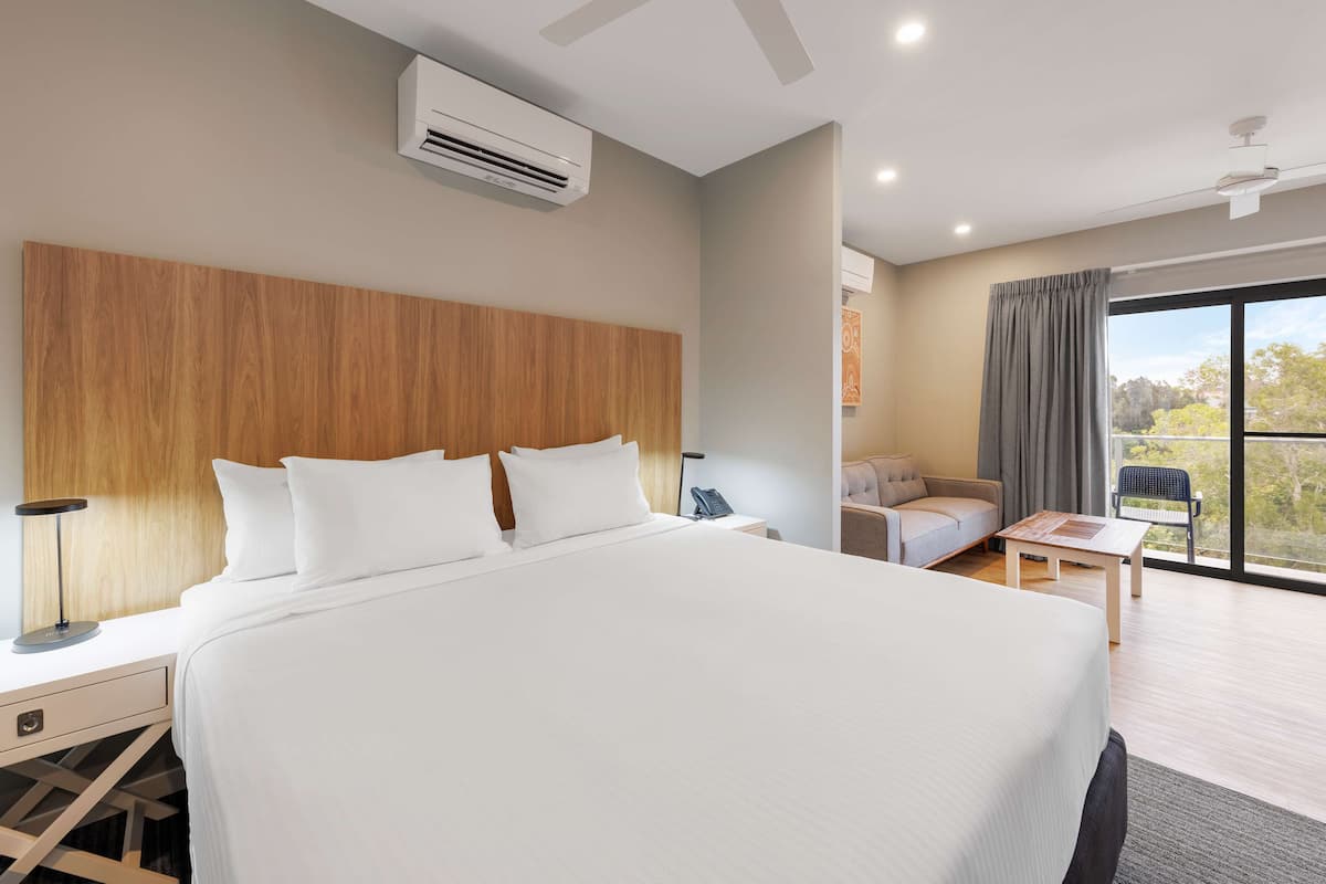 Une chambre d’hôtel moderne dotée d’un grand lit, d’une tête de lit en bois, d’un bureau avec une lampe, d’une petite table, d’un canapé et offrant une vue sur l’extérieur grâce à une grande fenêtre.