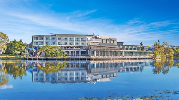Un edificio moderno con fachada de vidrio, un reflejo en el agua y un muelle con sombrillas.