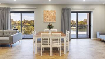 Una sala moderna con una mesa de comedor, sillas blancas, un sofá gris y un ventanal con vistas a árboles y un lago.