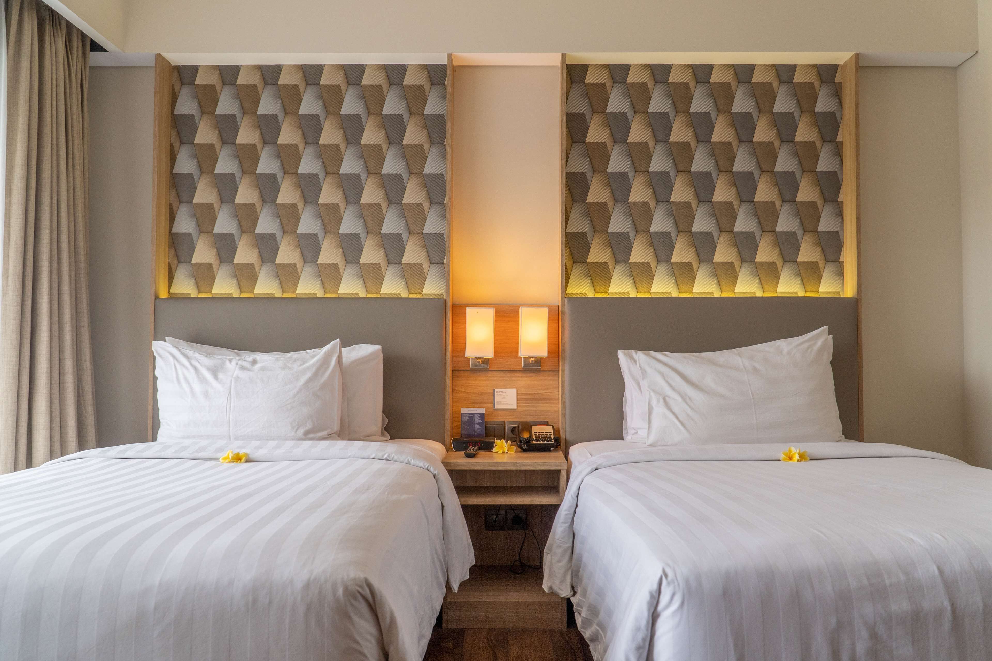 A hotel room with two beds, a wooden headboard with geometric patterns, a nightstand with a lamp, and a small table with a telephone and a book.