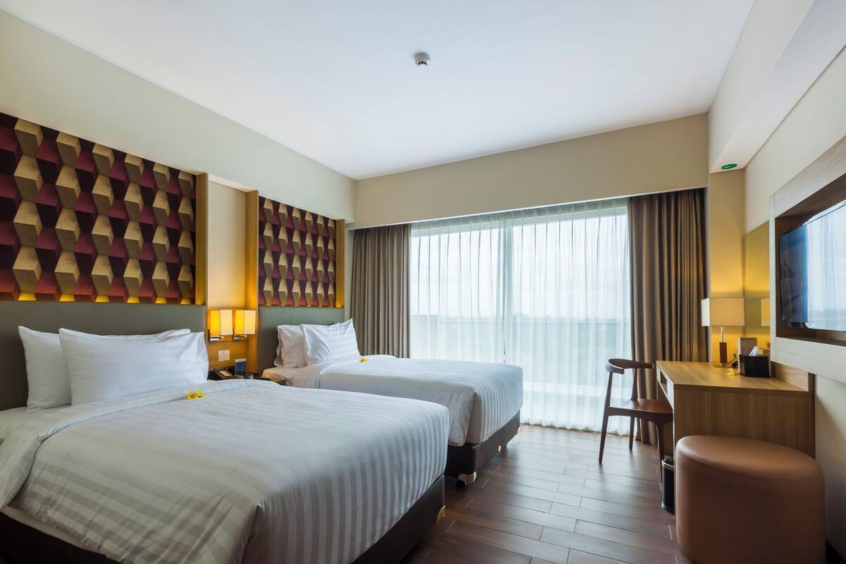 Habitació d'hotel amb dos llits, una taula de treball, una cadira i una finestra gran amb cortines.