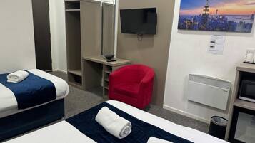 Habitación de hotel con dos camas, una silla roja, televisión, un cuadro de un paisaje urbano y una kitchenette.