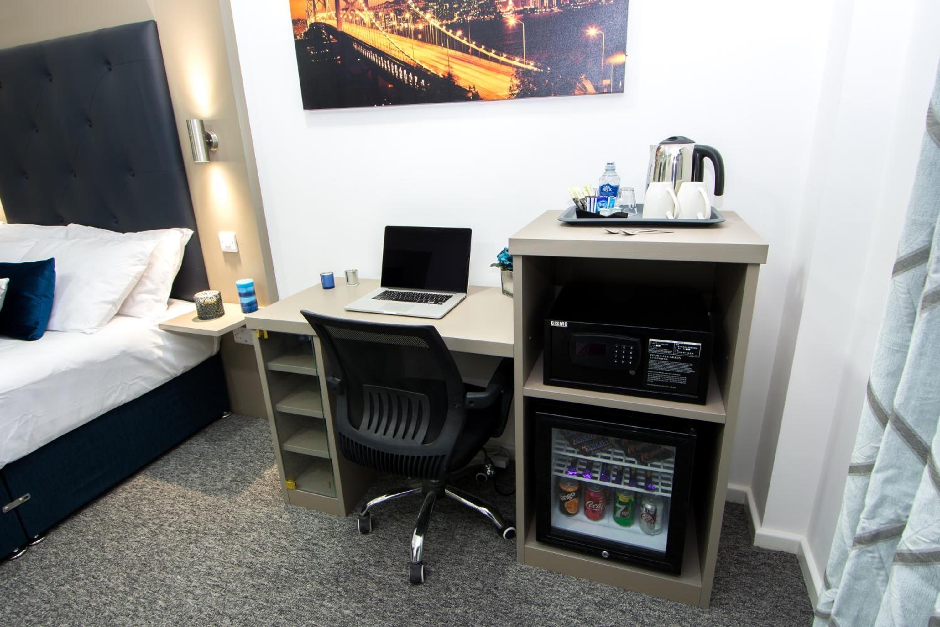 Habitación de hotel con cama, escritorio con laptop, silla, cafetera y minibar.