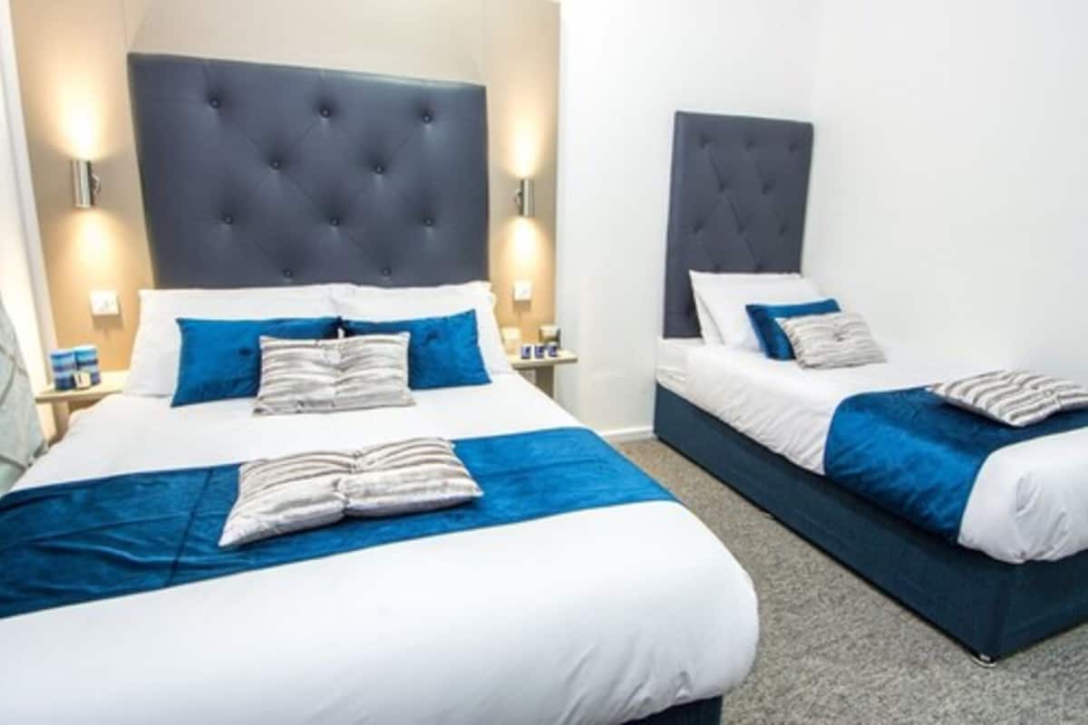 Ein Hotelzimmer mit zwei Betten, jeweils mit dunklem Kopfteil und weißer Bettwäsche. Zwei Nachttische mit Lampen stehen bereit, und das Zimmer ist in einer neutralen Farbgebung gehalten.