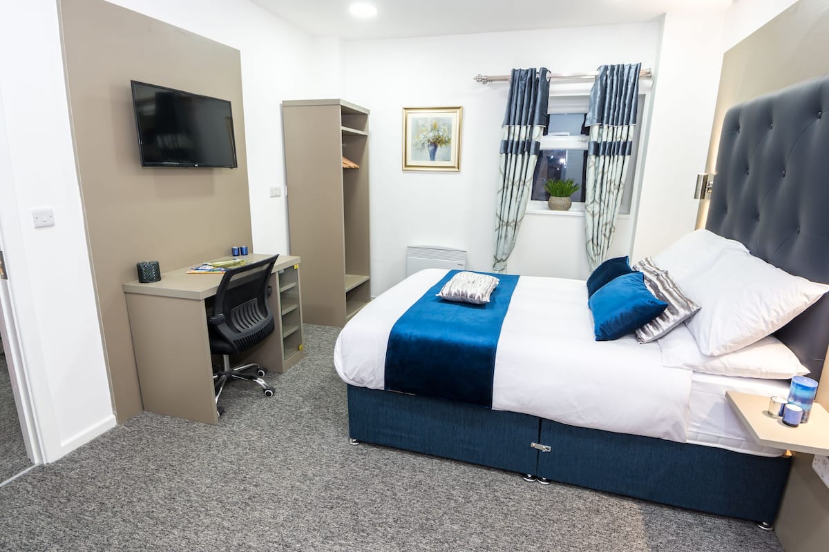Ein Schlafzimmer mit Bett, Schreibtisch, Stuhl, Fernseher, Kleiderschrank und einem Fenster mit Vorhängen.