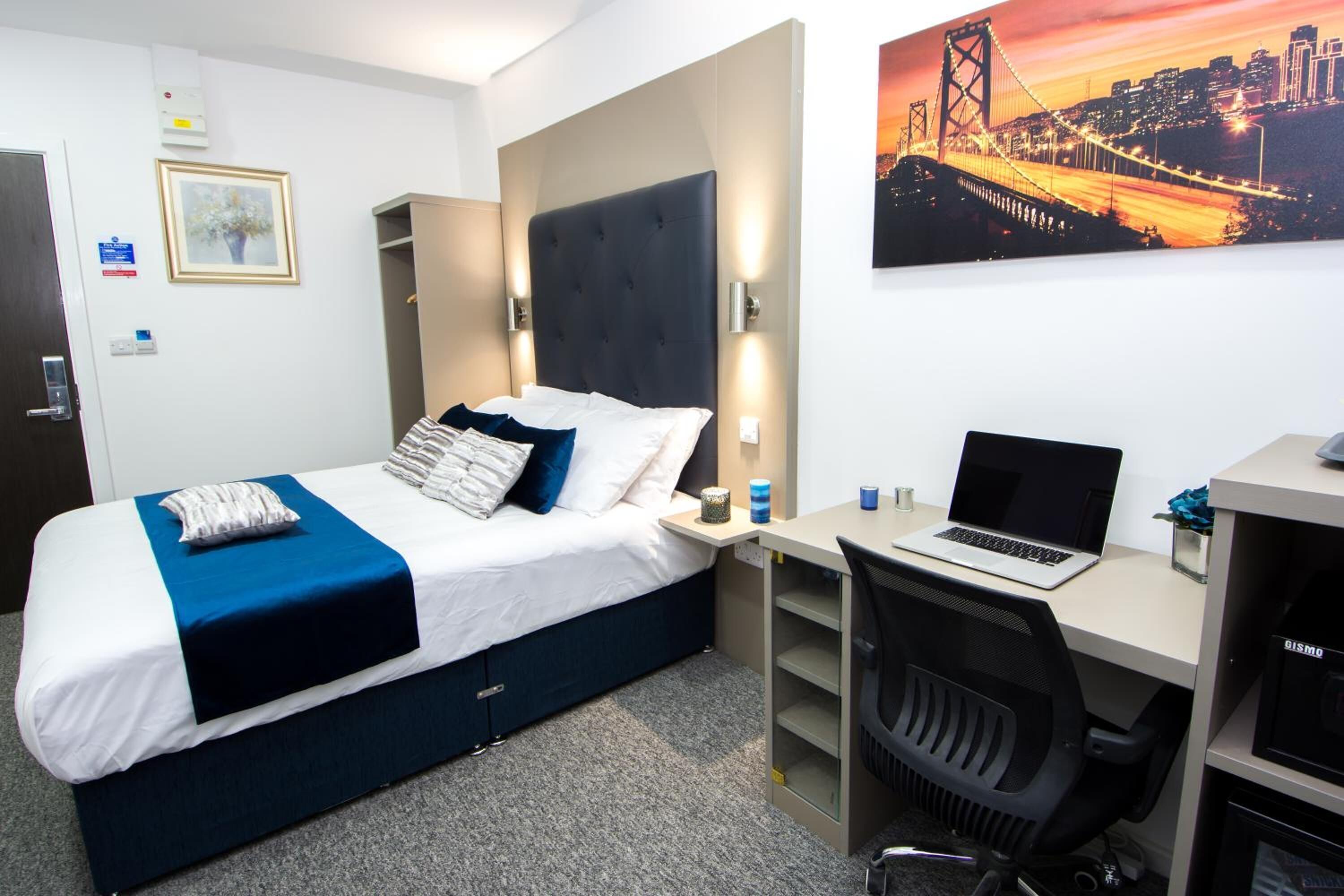 A hotel room with a bed, a desk with a laptop, a chair, and a cityscape painting on the wall.