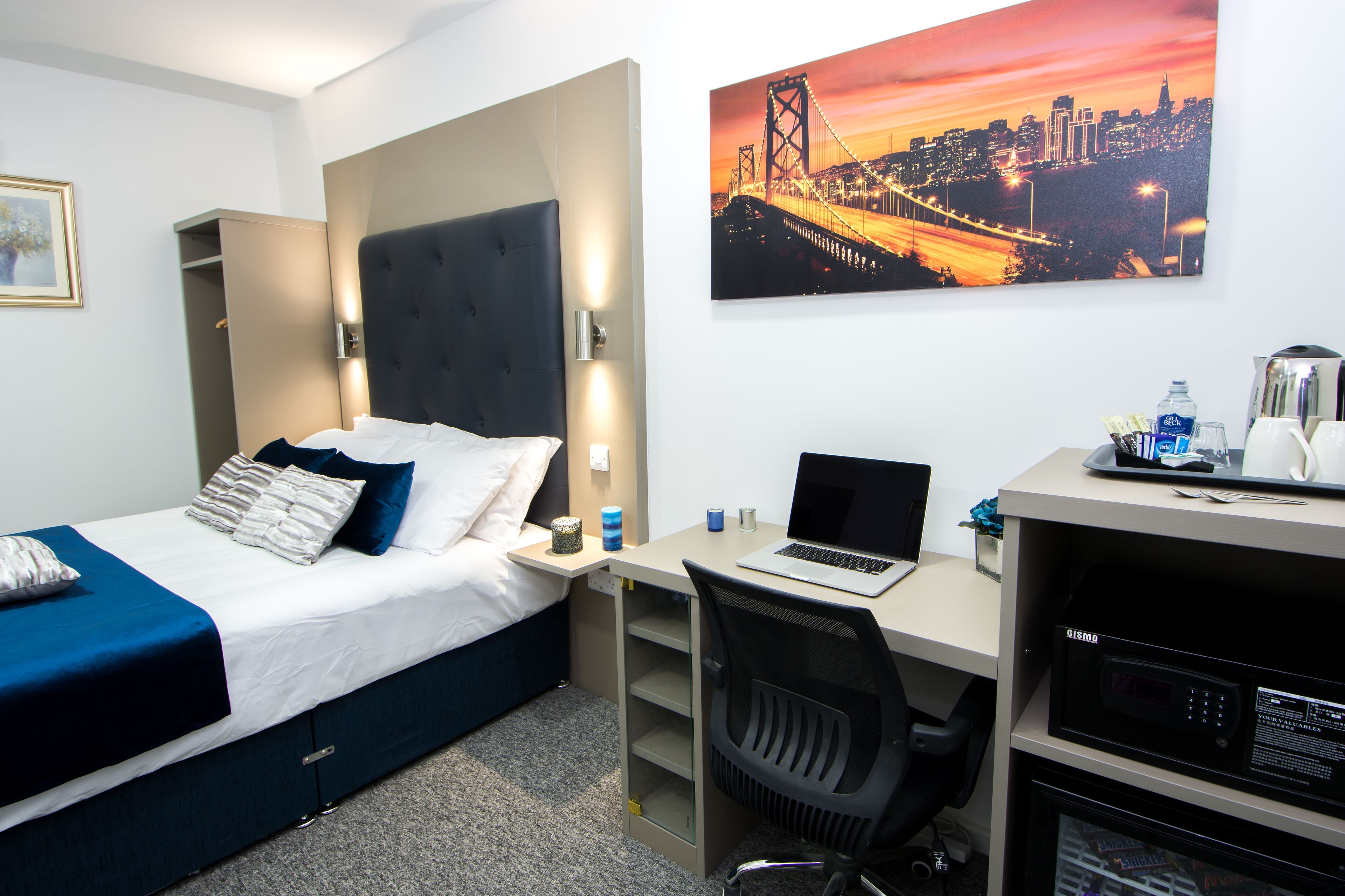 A hotel room with a bed, desk, and chair. There is a painting of a bridge on the wall.